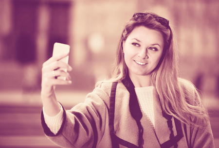 Young smiling glad cheerful woman looking curious and taking pictures with her phone outdoorsの写真素材