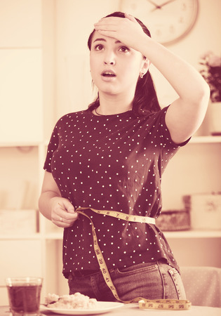 Girl measuring waistline with yellow tape frustrated by increasing in volumeの写真素材