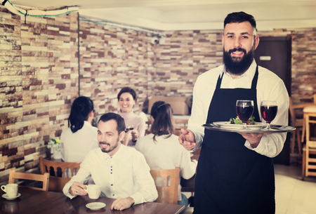 Cheerful male waiter serving rural restaurant guests at tableの写真素材