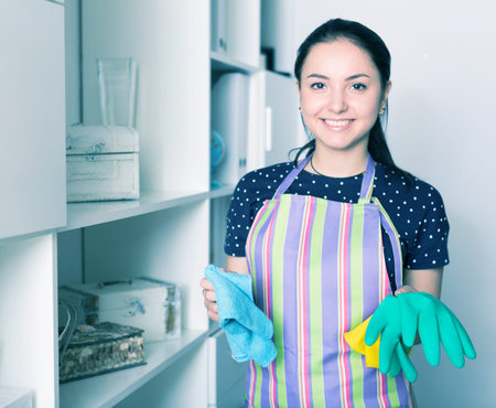 Cheerful young woman wiping shelves with rubber gloves in handの写真素材