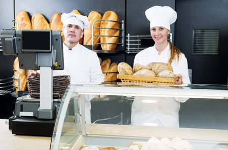 Happy male and young female staff offering fresh baguettes and buns in bakery