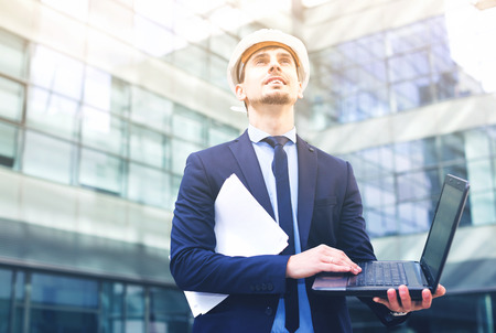Smiling man with documents in suit and helmet working at the laptopの写真素材