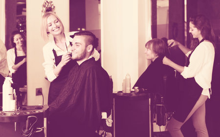 Smiling professional young hairdresser doing hairstyle for young men