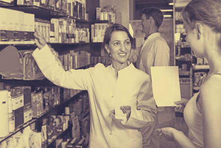 Positive woman pharmacist in white coat helping customers to find item in drug storeの写真素材