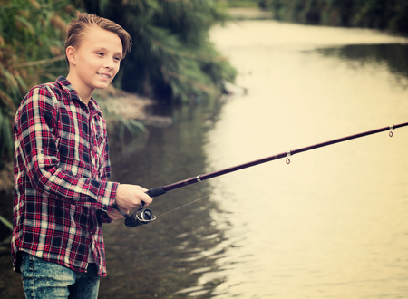 Positive teenager boy fishing using rod from water side on riverの写真素材