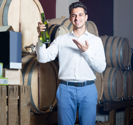 smiling man choosing bottle of white wine in shop with woodsの写真素材