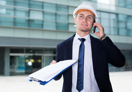 Foreman in suit and hat with folder is taking folder and discussing project by the phone near the building.の写真素材