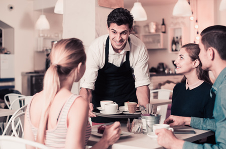 Polite happy waiter bringing ordered dishes to friends in tearoom of a cozy confectioner's shopの写真素材
