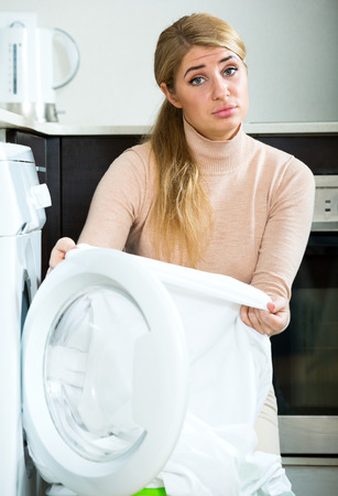 Unhappy blonde woman with dirty white shirt near washing machineの写真素材