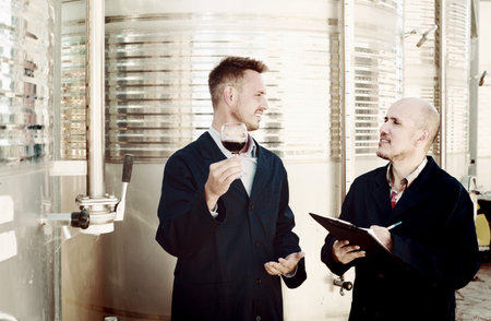 Two male employees checking  wine sample in glass and taking notes on winery manufactory