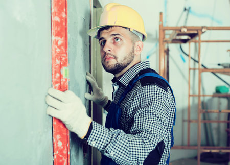 Builder in helmet is measuring wall with level.