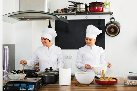 Cheerful professional mature and young women cook wearing uniform standing on kitchenの写真素材