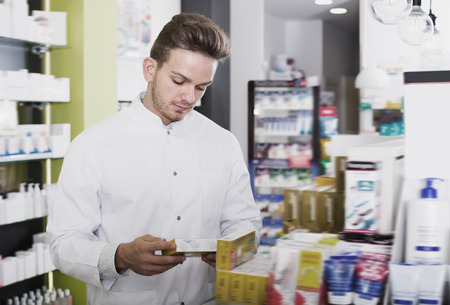 Glad man pharmacist wearing white coat standing among shelves in drug storeの写真素材