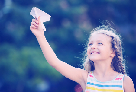 smiling girl in bright dress playing with flying paper airplane toy outdoorsの写真素材