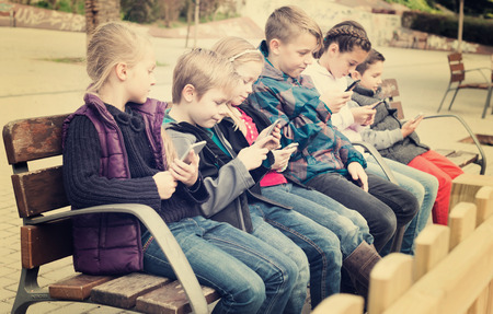 Preschool children sitting on bench with mobile devices outdoorの写真素材