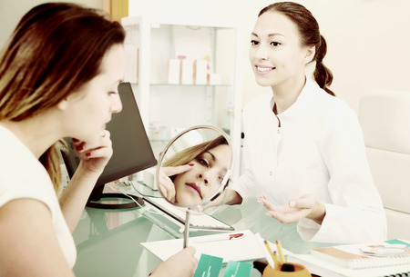 Young woman looking at mirror during consultation with beautician in aesthetic medicine centerの写真素材