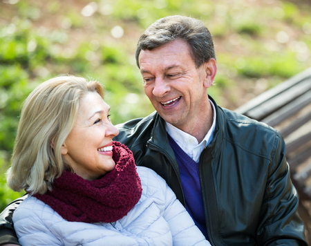 Portrait of smiling aged couple enjoying spring day on the bench in park . Focus on manの写真素材