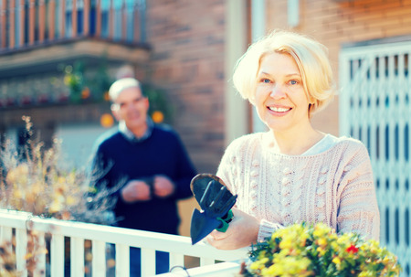 Senior woman with gardening accessories on a terrace near her house with her husband holding a cup of coffee in a backgroundの写真素材