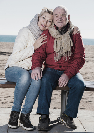 Aged husband and wife sitting together on bench by sea on chilly dayの写真素材