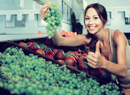 positive young russian woman customer choosing grapes in fruit storeの写真素材