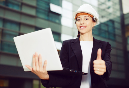 Smiling female designer in suit and hat with laptop is pleased with realisation her project on the outside.の写真素材