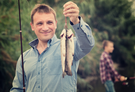 portrait of happy man showing fresh catch fish on lake shoreの写真素材