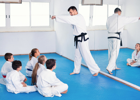 Photo for Smiling young cheerful coach showing new martial moves to children in karate class - Royalty Free Image