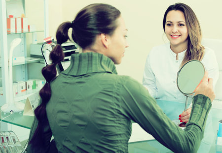 Young smiling woman looking at mirror during consultation in aesthetic medicine centerの写真素材