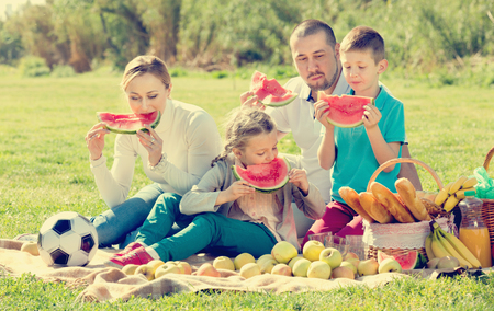 Cheerful positive family of four having a picnic and eating watermelon outdoors in a sunny weatherの写真素材