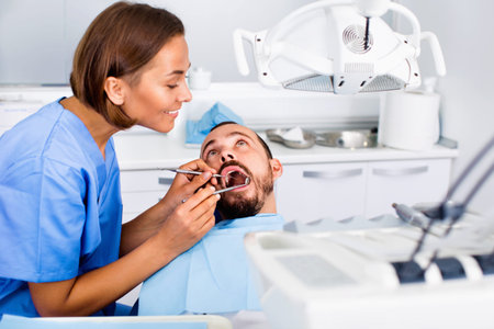 Positive smiling stomatologist girl in uniform is taking visional inspection of a man on the chair in dental clinic.の写真素材
