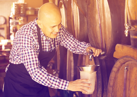 Elderly man wine maker taking wine from wood in wineryの写真素材