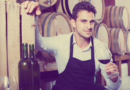 Glad man seller in apron holding glass of red wine in shop with wooden barrelsの写真素材