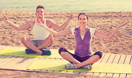 Charming smiling woman and man sitting cross-legged do yoga poses on beach at daytimeの写真素材