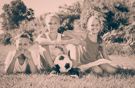 Three cheerful positive  kids with ball resting on grass on sunny summer dayの写真素材