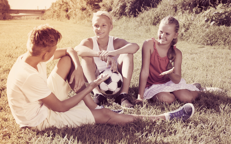 girl with two happy friends resting after game with ball on green grassの写真素材