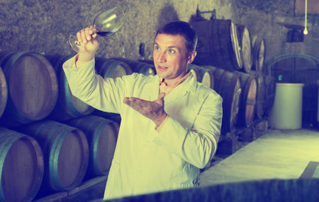 Young sommelier holding glass of red wine and checking quality of red wine in barrels