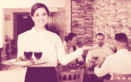 Smiling woman waiter carrying order for visitors in country restaurantの写真素材