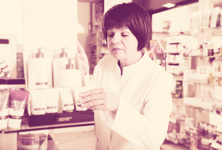 Glad woman druggist wearing white coat standing among shelves in drugstoreの写真素材