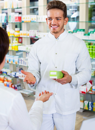 Positive man pharmacist wearing uniform assisting customers in pharmaceutical shopの写真素材