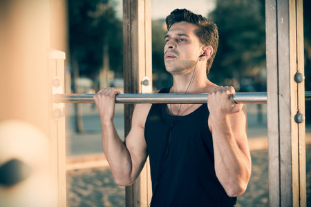 Sporty man enjoying music during workout outdoors, doing chin-ups on horizontal bars