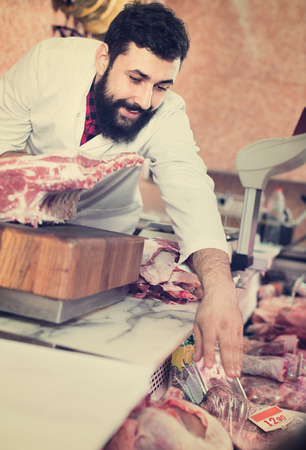 Smiling cheerful positive man seller grouping meat to sell in butchers shopの写真素材