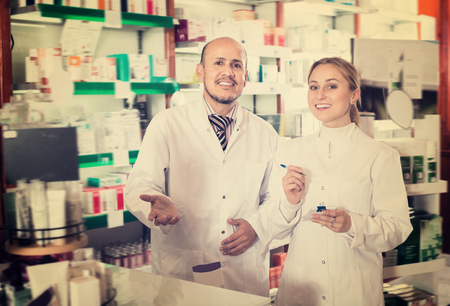 Two smiling pharmacists wearing white coats standing next to shelves with medicineの写真素材
