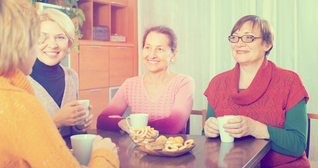 Positive smiling female pensioners drinking coffee and talking at homeの写真素材