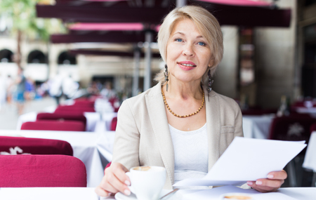 Mature businesswoman is reading documents in time breakfast in cafe.の写真素材