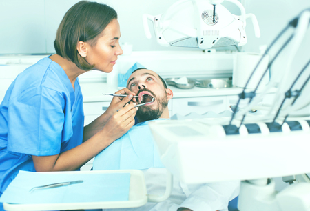 Smiling stomatologist girl in uniform is taking visional inspection of a man on the chair in dental clinic.の写真素材