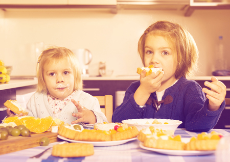 Two happy little sisters enjoying pastry with cream in kitchenの写真素材