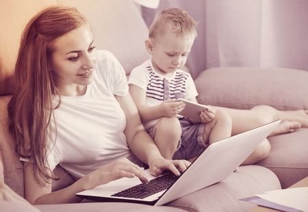 Satisfied mother is productively working behind laptop while kid playing on tablet at home.の写真素材