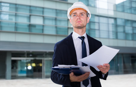 Engineer in suit and hat with folder is looking into documents with project near the building.