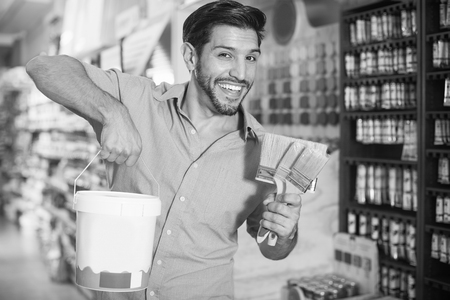 Smiling male buyer holding bucket of paint and brushes in paint storeの写真素材