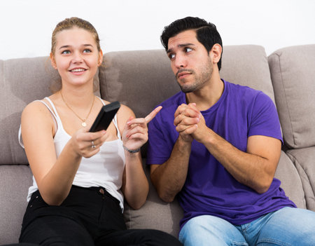 Guy sitting on sofa begging his girlfriend to give him TV remote control